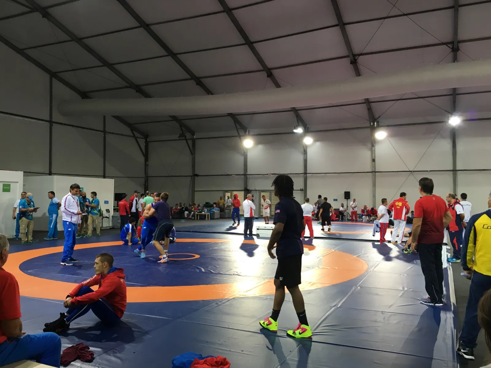 Wrestlers and coaches from several national teams warming up on the mats of a competition training hall.